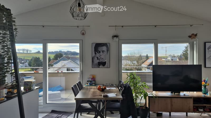 Bright living space with large windows offering a scenic view, featuring a dining table with chairs, a TV screen, potted plants, and artwork on the walls.