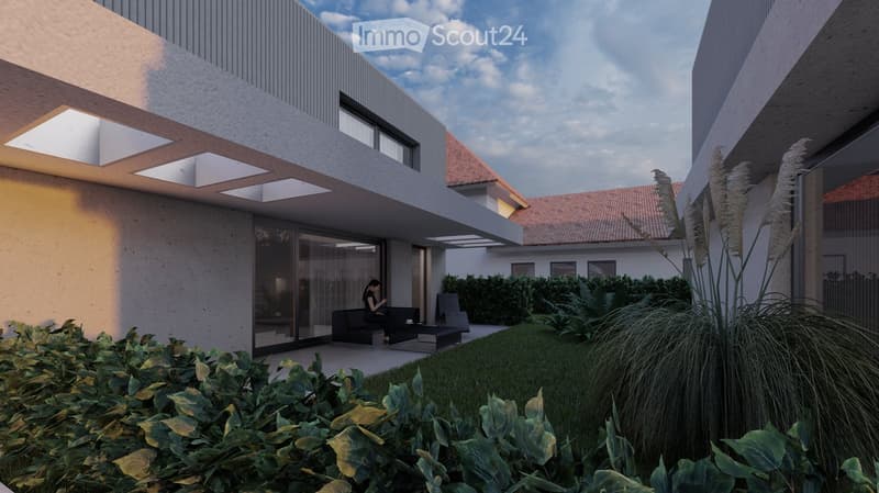 Two-story modern house with a gray exterior, large windows, and a covered patio area. The house is surrounded by lush greenery, including shrubs and tall grasses.