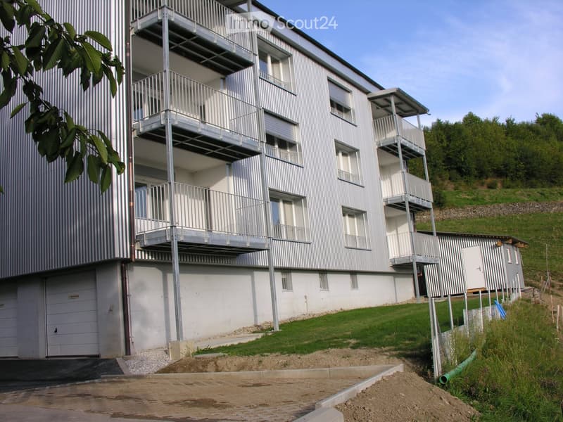 Modern multi-story building with white and grey exterior, multiple balconies, garage, and surrounding greenery.