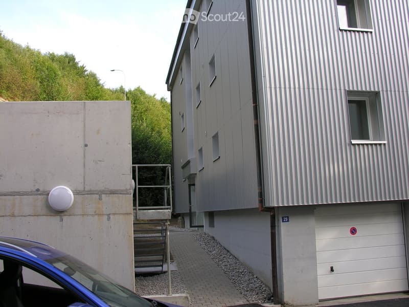 Modern two-story building with a metallic facade, concrete wall, and garage entrance. A raised ground floor leads to the main entrance.