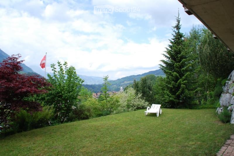 Lush green garden with a white lounge chair, surrounded by trees and a scenic mountain view.