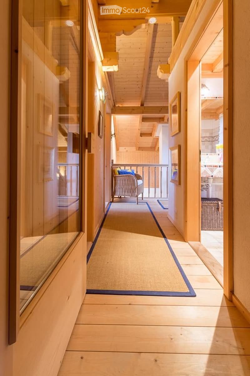 A well-lit hallway with wooden floors, exposed wooden beams, and a carpeted area leading to a balcony. The hallway features glass-paneled doors and framed artwork on the walls.