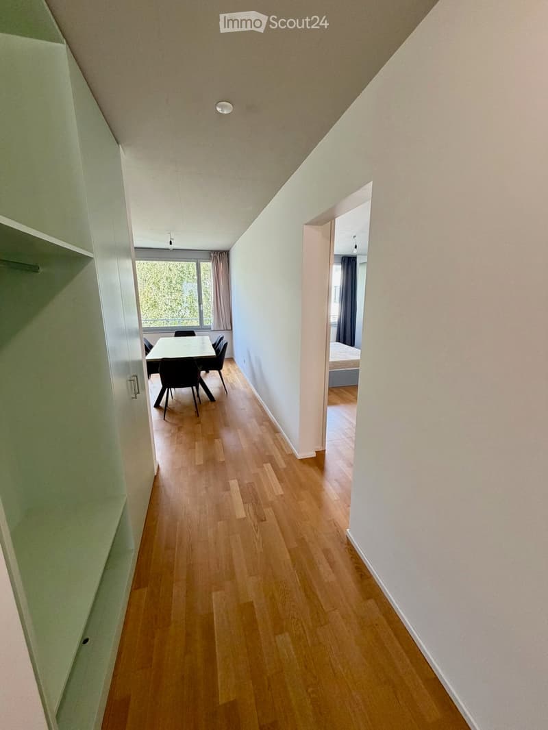 A narrow hallway with wooden flooring, leading to a dining area and a bedroom. The hallway has white walls and a ceiling light fixture.