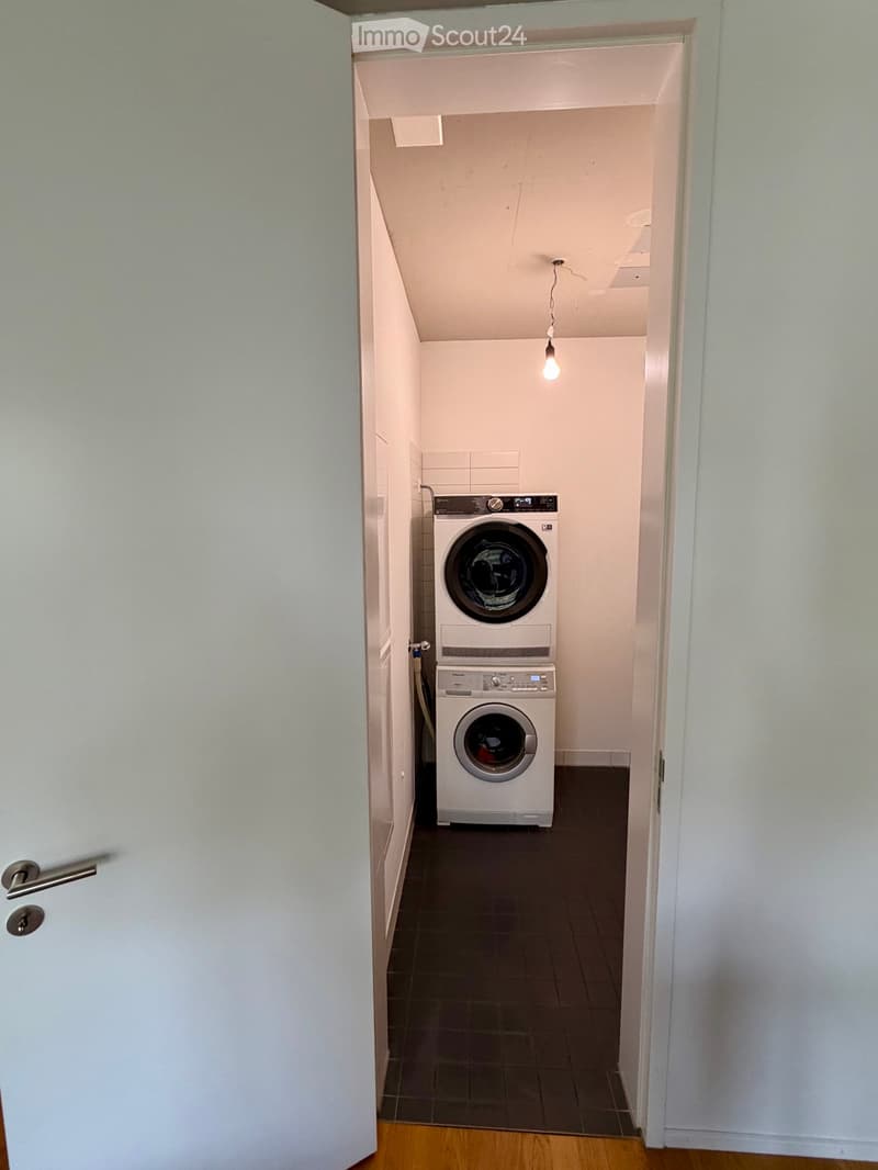 A small, well-lit laundry room with a washing machine and dryer stacked vertically, featuring tiled walls and a dark tiled floor.