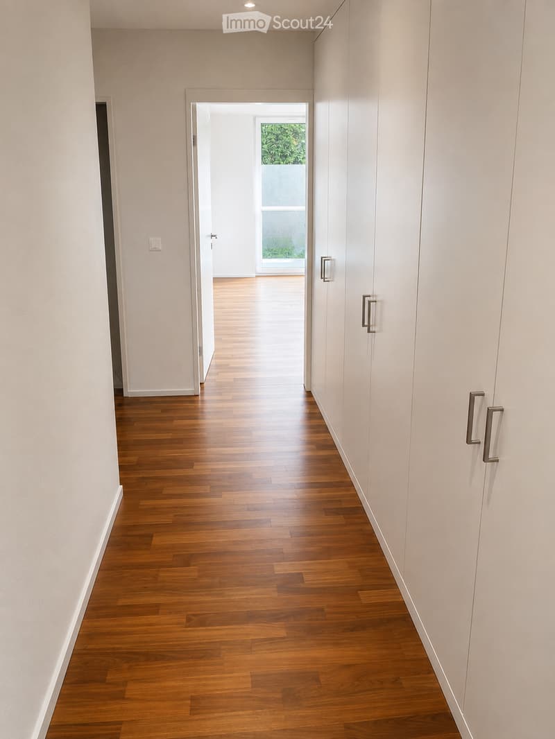 A clean, modern hallway with light-colored wooden flooring and white walls, featuring built-in storage units with sleek metal handles and a recessed ceiling light.