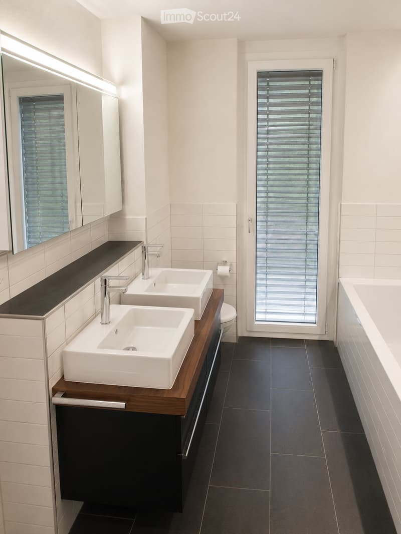 Modern bathroom with two white sinks on a dark wooden vanity, chrome faucets, a large mirror, a toilet, and a bathtub. The room features white tiled walls, dark tiled flooring, and a window with blinds.