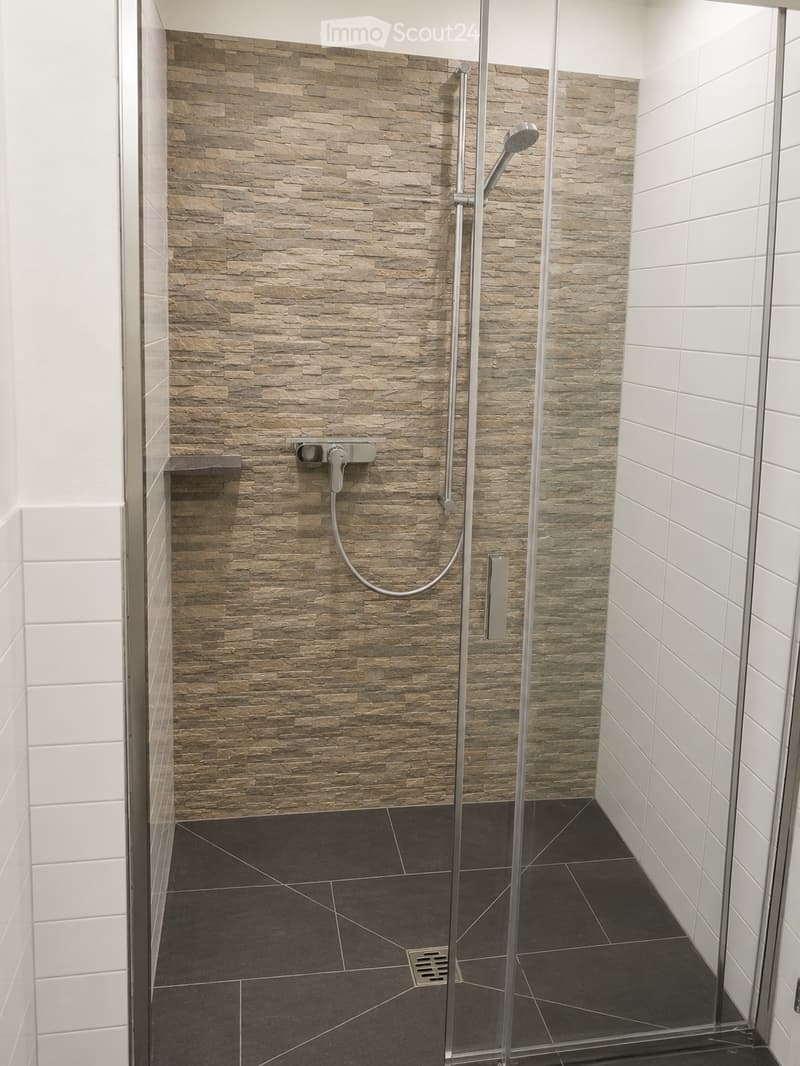 Modern bathroom with a glass-enclosed shower area, stone-textured wall tiles, and dark tiled flooring.