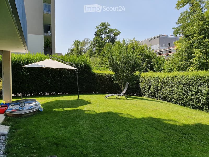 Well-maintained lawn with a large umbrella, sun lounger, and surrounding greenery, including bushes and trees. Part of a residential building is visible on the left.
