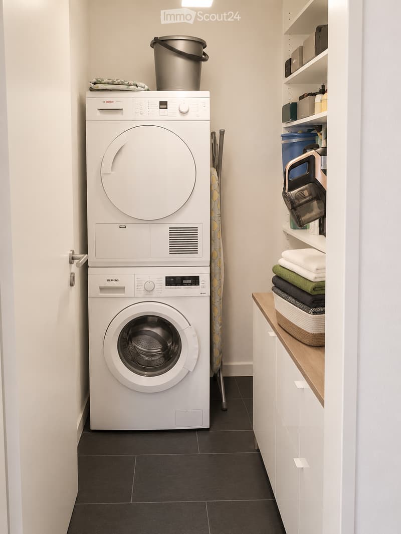 A compact bathroom with a stacked washing machine and dryer, shelves with cleaning supplies, and a small wooden shelf holding towels and a basket.