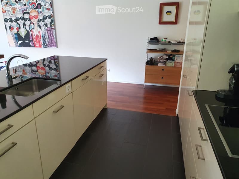Modern kitchen with light-colored cabinets, dark countertops, a stainless steel sink, and a coffee maker on the counter. The floor is dark tiled, and there is a small wooden drawer unit with various items on top.
