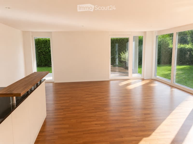 Empty room with wooden flooring, white walls, and large windows overlooking a green garden. A wooden counter is visible on the left side.