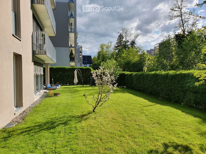 Well-maintained garden with green lawn, small tree, hedge border, and modern apartment building in the background.