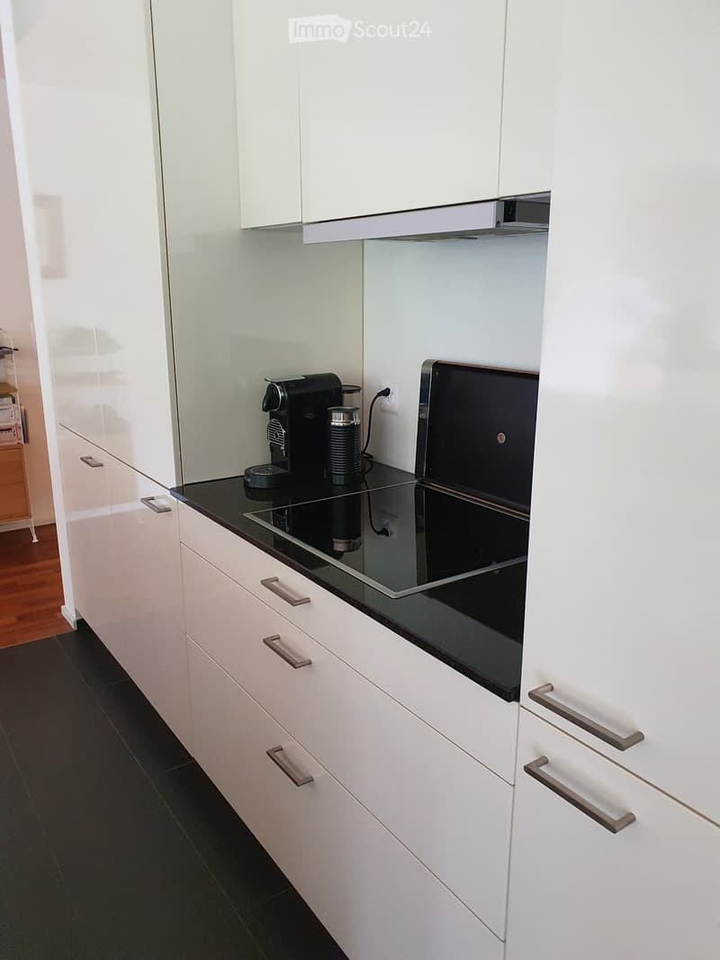 Modern kitchen with white cabinets, black countertop, and stainless steel handles. Features a coffee maker and an induction stove.