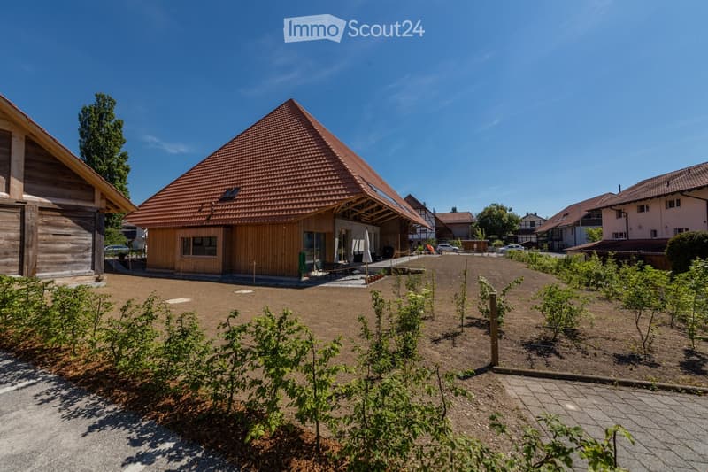 A modern wooden house with a triangular roof and large windows, surrounded by a landscaped garden area with young plants and a gravel pathway.