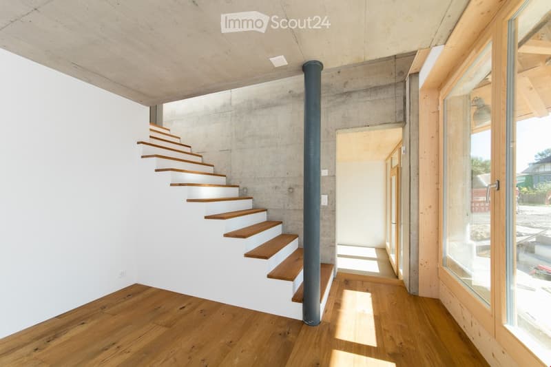 Modern hallway with wooden flooring, white concrete walls, and a staircase with wooden steps leading upwards. Features large windows and a central black column.