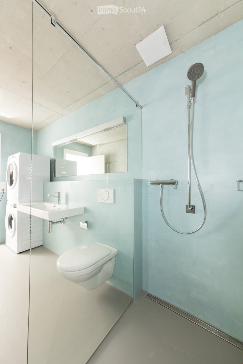 Modern bathroom with light blue walls, white fixtures, glass shower enclosure, and built-in washing machine.