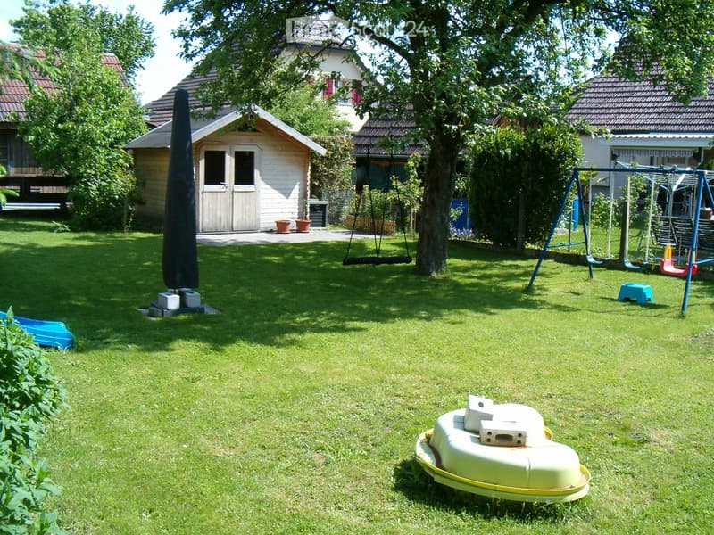Well-maintained lawn with a swing, slide, and small play structure. A shed and patio umbrella are visible in the background.