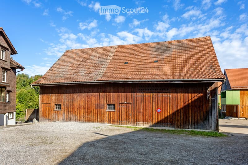 A traditional wooden barn with a red-tiled roof, large wooden doors, and small windows, situated in a rural area with gravel ground and surrounding greenery.