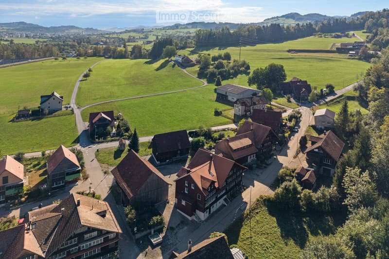 Aerial view of a rural village with traditional houses, surrounded by green fields and trees, with a road winding through the area.