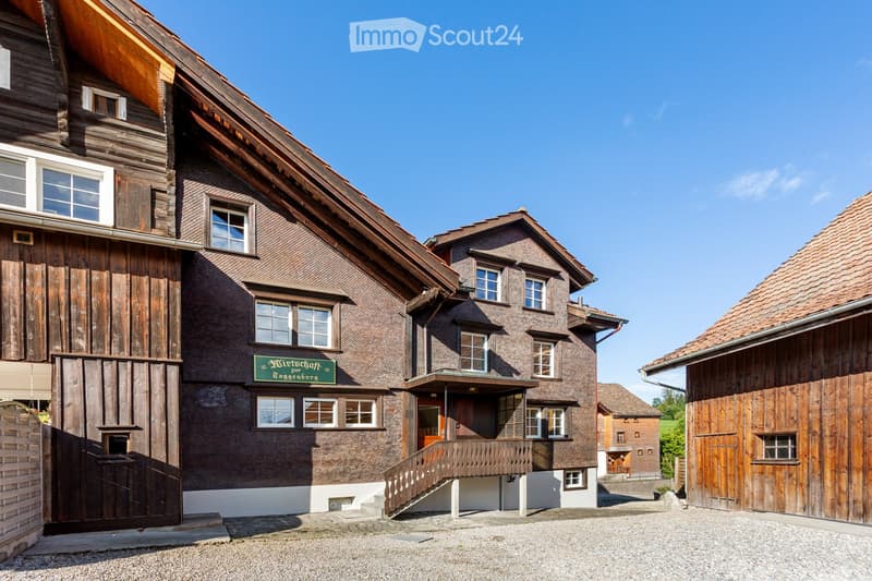 Traditional wooden house with a steep roof, multiple windows, and a sign reading "Wirtschaft Zogenburg" on the facade. The building is surrounded by other wooden structures and a gravel driveway.