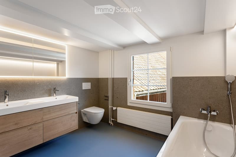 Modern bathroom with a double sink, toilet, bathtub, large window, and built-in storage cabinet.