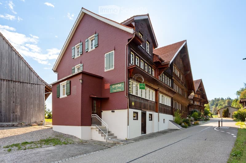 A two-story building with red brick walls and green shutters, featuring a wooden balcony and a sign reading "Mietshaus zur Gögenburg".