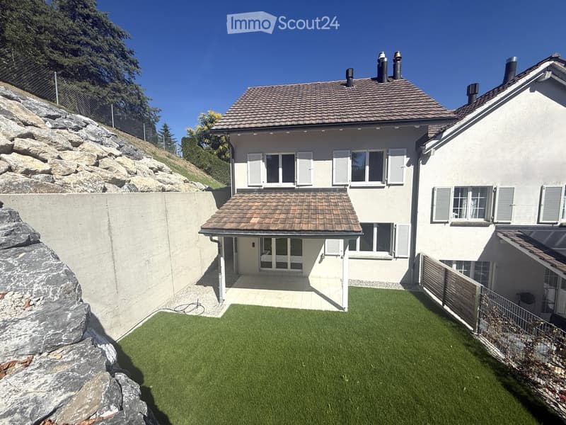 Two-story house with a tiled roof, multiple windows with shutters, and a small attached garage. The property includes a well-maintained lawn and a stone retaining wall.