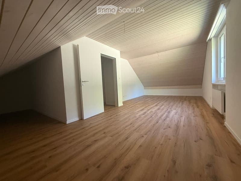 Empty attic room with wooden flooring, white walls, and a slanted wooden ceiling. Features a single window and a closed door leading to another space.