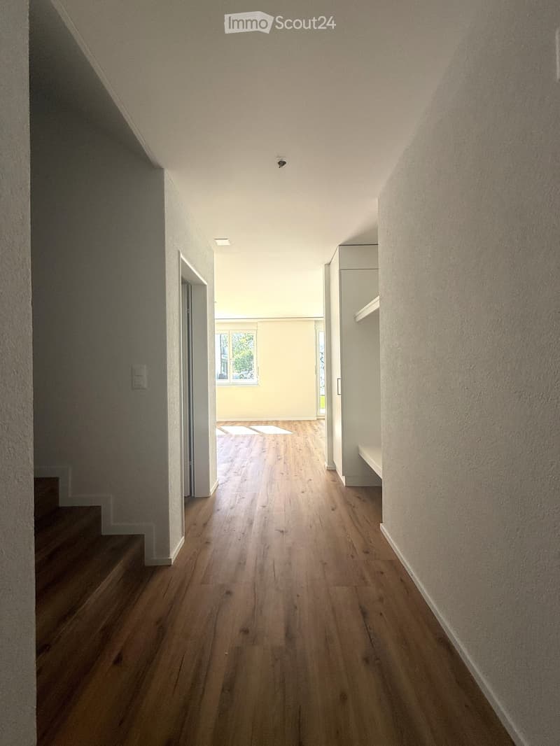 A modern hallway with wooden flooring, white walls, and recessed lighting. The hallway leads to a room with a window and a door.