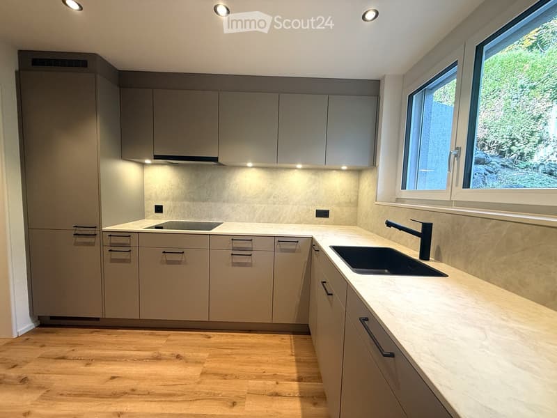 Modern kitchen with light grey cabinets, a central island, and a sink with a modern faucet. The countertops are light-colored, and the floor is wooden.