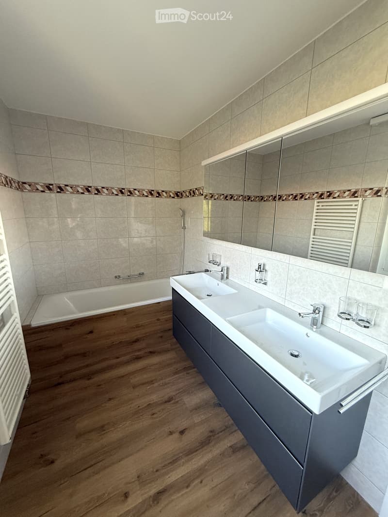 Modern bathroom with double sink, large mirror, bathtub, and wooden flooring.