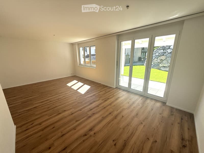 Empty room with wooden flooring, large sliding glass doors leading to a garden, and a window on the left side.
