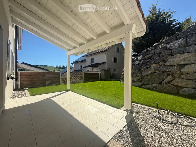 A covered patio area with tiled flooring, leading to a well-maintained lawn surrounded by a stone wall and wooden fence. A house is visible in the background.