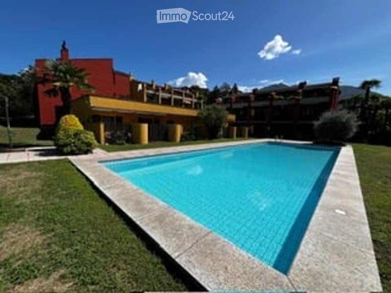 Outdoor area with a swimming pool, surrounded by grass and a few plants. The background shows a building with a red and yellow facade and a clear blue sky.