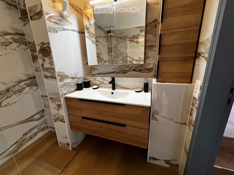 Modern bathroom with a white sink, wooden vanity, marble-patterned tiles, and a large mirror above the sink.