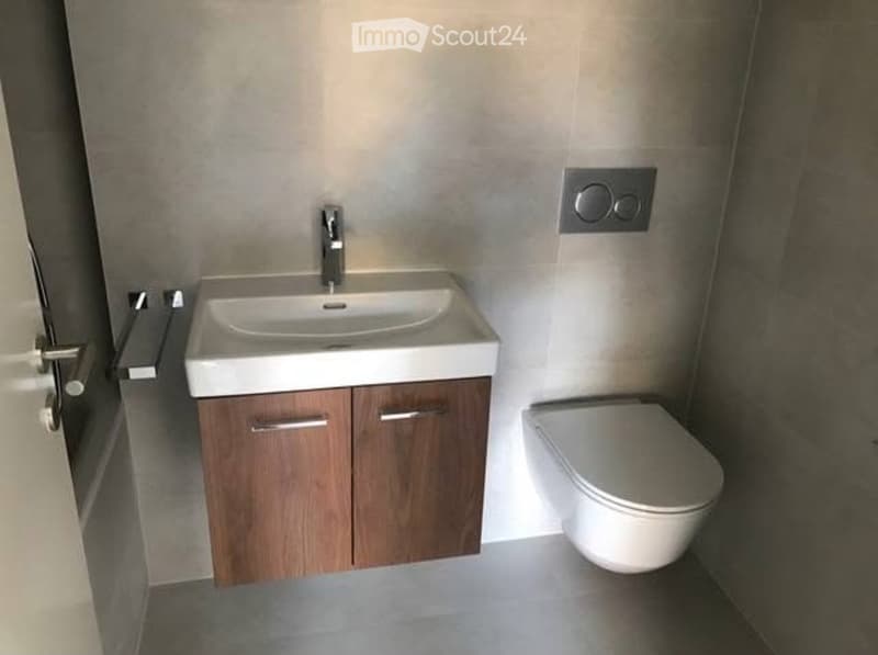 Modern bathroom with a white sink, wooden cabinet, and toilet. The walls are tiled in a light grey color, and the floor is a smooth grey surface.