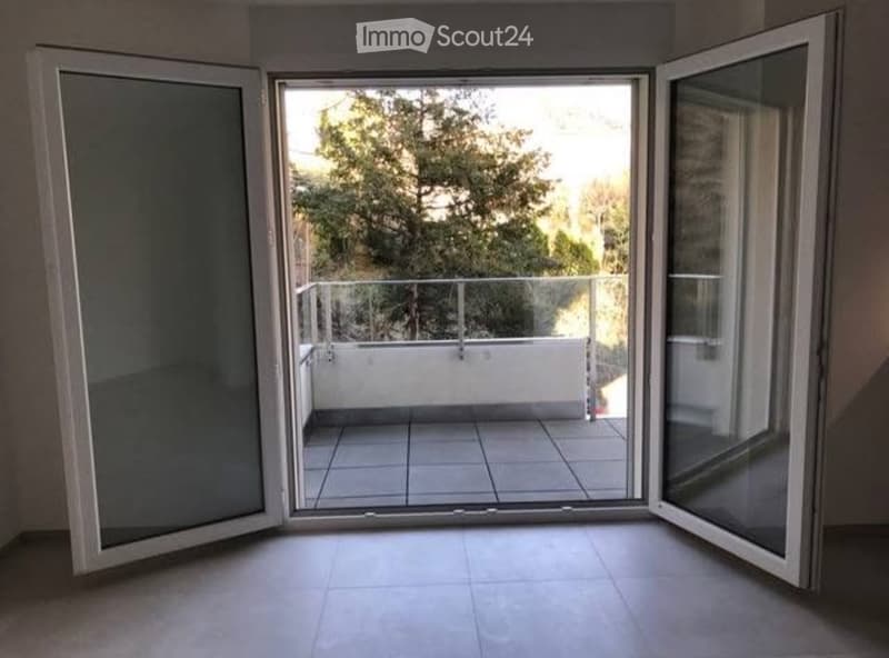 Open sliding doors lead to a tiled balcony with a glass railing, overlooking a tree-filled outdoor area.
