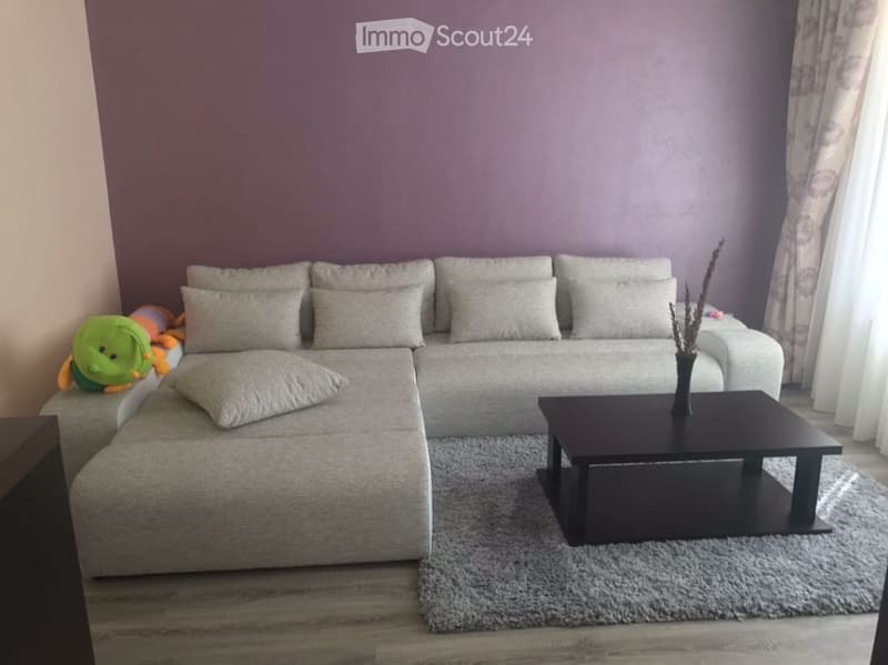 Modern living room with a grey sectional sofa, multiple pillows, a black coffee table with a vase, and a plush toy on the sofa. The room features wooden flooring and large windows with curtains.