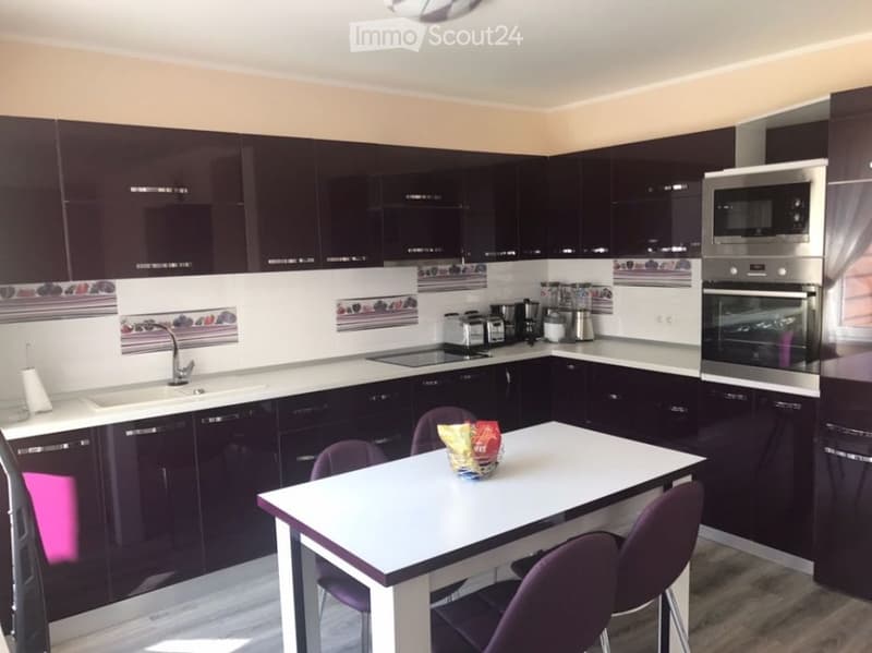Modern kitchen with dark cabinets, white countertops, and stainless steel appliances. Features a sink, microwave, oven, toaster, and a small dining area with a table and chairs.