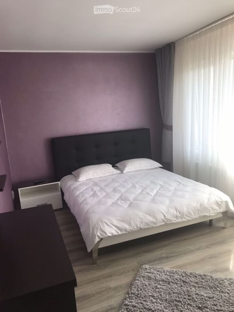 Modern bedroom with a large bed, white bedding, nightstand, and a grey rug on the floor. The walls are painted in a soft purple color, and there are dark curtains on the windows.