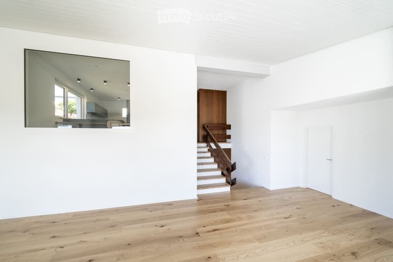 Bright, minimalist hallway with white walls, wooden flooring, and a staircase leading upwards. A large mirror reflects part of the adjacent kitchen area.