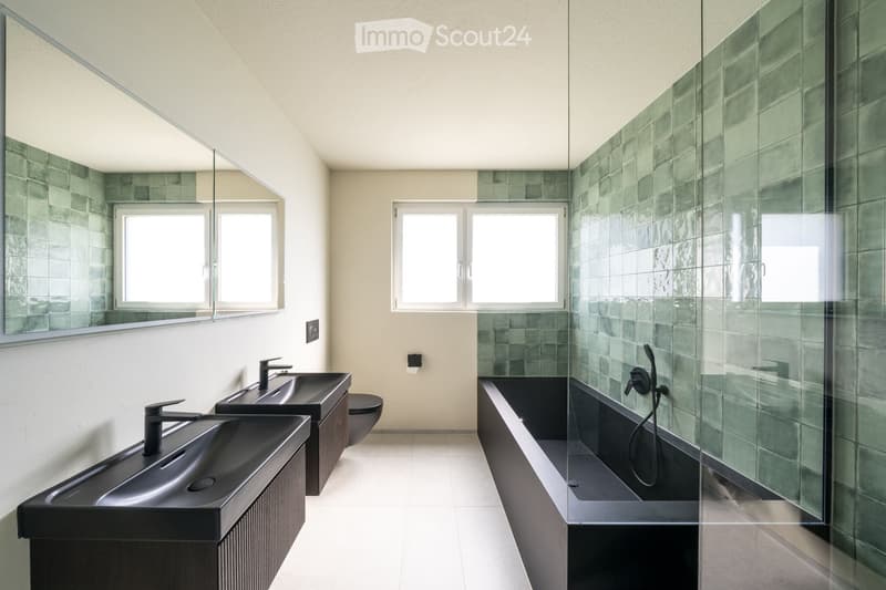 Modern bathroom with two black sinks, a large mirror, a bathtub with glass enclosure, and a toilet. Green tiled walls and white flooring.