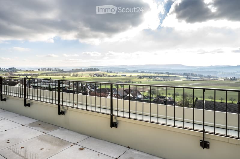 Balcony with metal railing, tiled floor, overlooking a rural landscape with fields, houses, and distant hills.