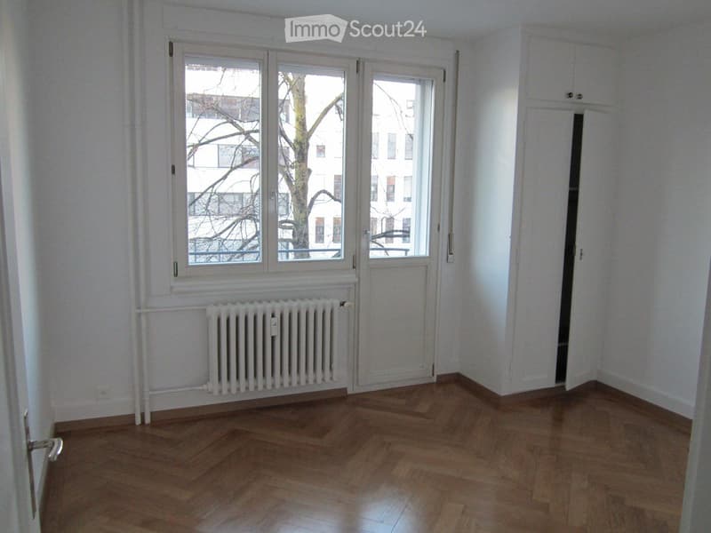 Empty room with wooden flooring, white walls, large windows, radiator, and a door leading to another space.