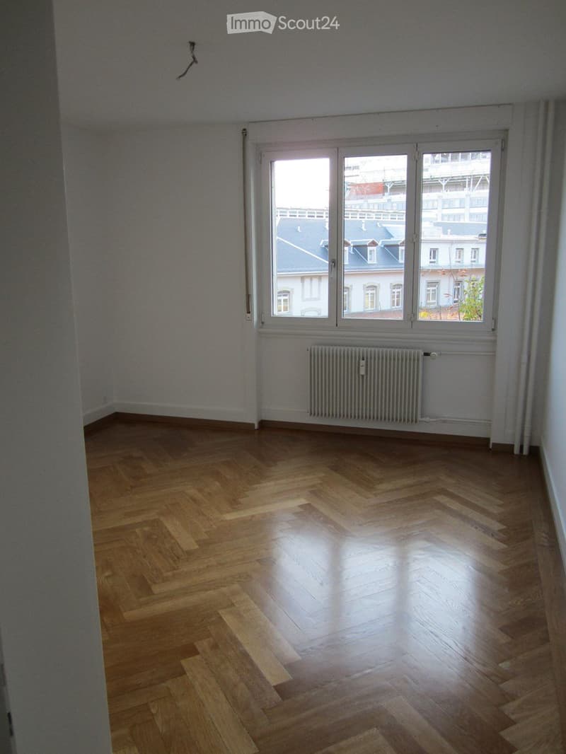 Empty room with wooden herringbone flooring, large window with white frame, radiator below window, and white walls.