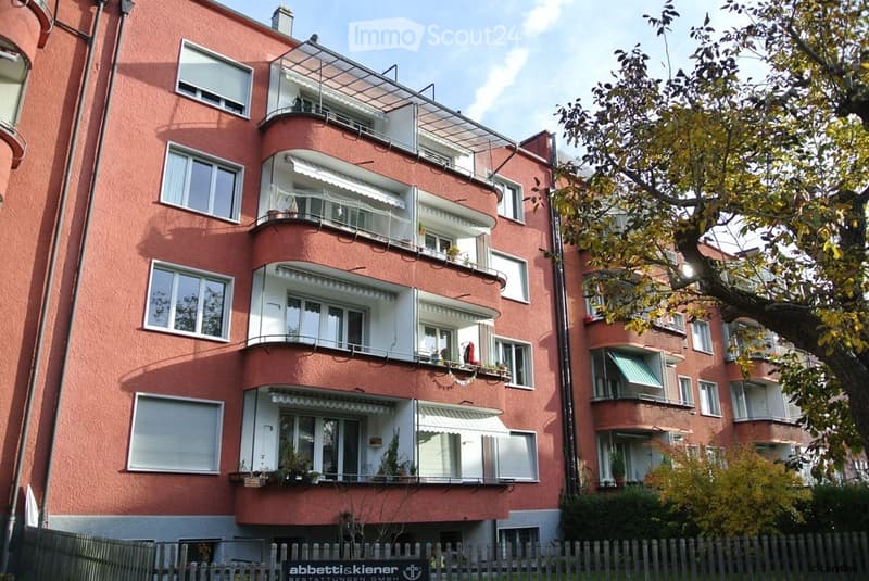 Four-story red brick apartment building with multiple balconies and awnings, surrounded by trees and a wooden fence.