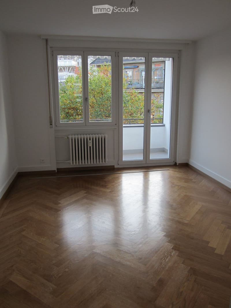 Empty room with wooden herringbone flooring, large sliding glass doors leading to a balcony, and a radiator under the window.