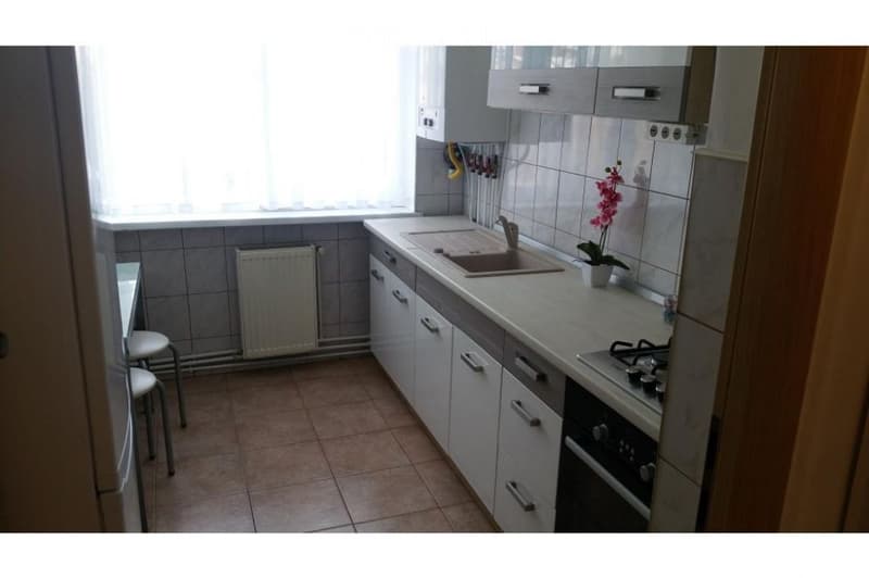 A compact kitchen with white cabinets, a sink, stove, and a small stool near the window. The space features tiled walls and a tiled floor, with a radiator under the window and a flower pot on the counter.