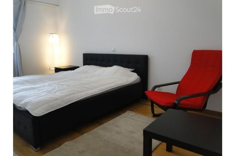 A modern bedroom with a large bed, a red chair, a small table, a nightstand, and a curtain on the window.