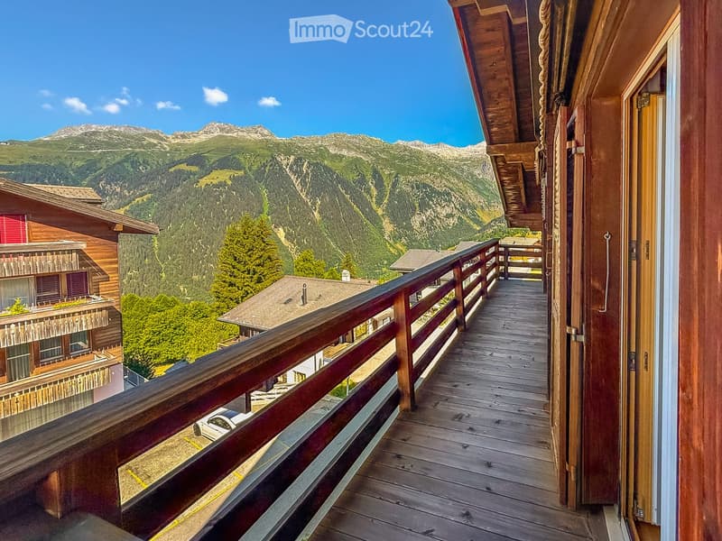 Wooden balcony with railing, overlooking a scenic mountain landscape with trees and neighboring houses.